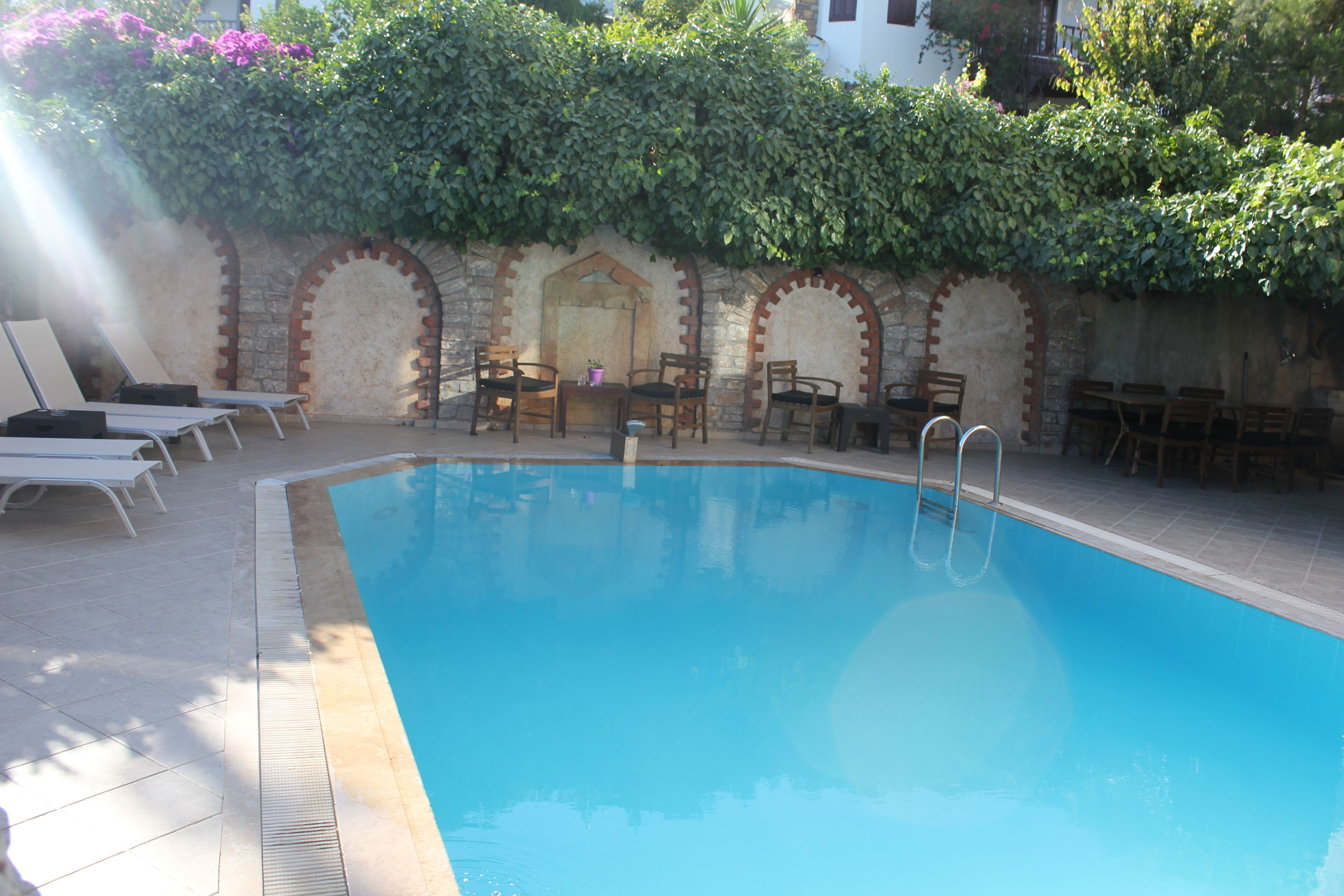 Una piscina con sillas y mesas de descanso, rodeada por un muro de piedra y follaje.
