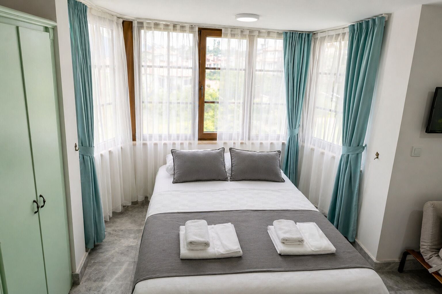 Habitación de hotel con cama, dos almohadas, ventana con cortinas, radiador, televisor y una silla.