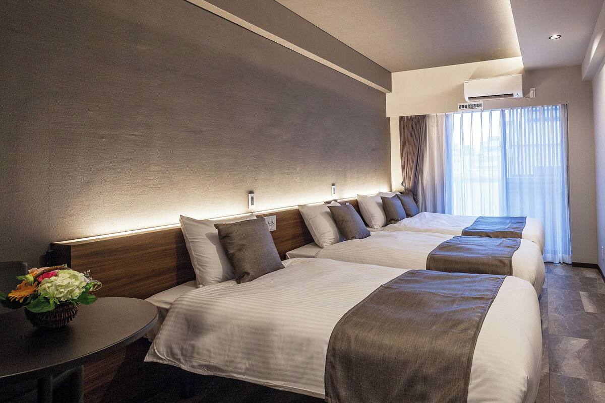 Une chambre d’hôtel au design moderne, comprenant un grand lit avec une tête de lit, une table de chevet agrémentée d’un vase de fleurs et une grande fenêtre avec des rideaux.