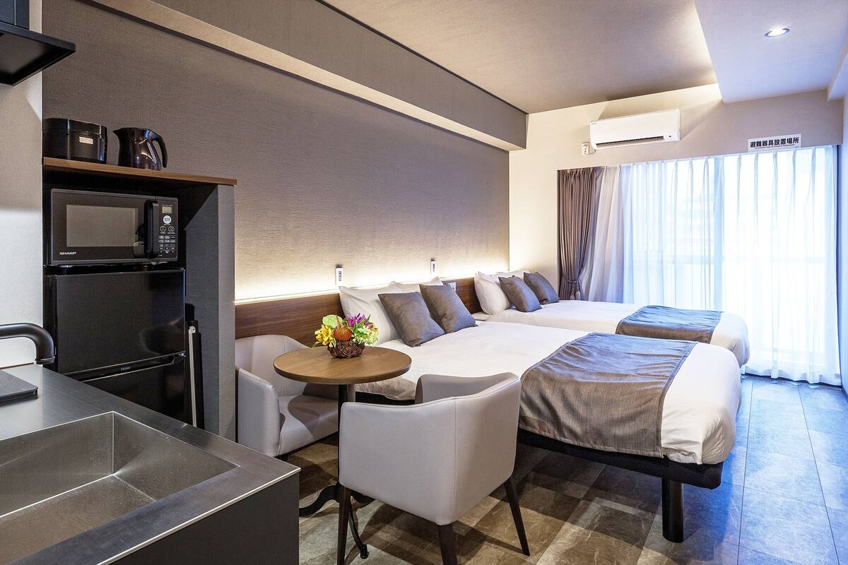 Une chambre d’hôtel moderne équipée d’une kitchenette, d’un canapé-lit, d’une petite table avec un panier de fruits et d’une grande fenêtre avec des rideaux.