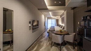 Una habitación de hotel moderna con un largo pasillo, un área de comedor con una mesa redonda y sillas, una kitchenette con microondas, y una cama con televisor y televisor de pantalla plana montado en la pared.