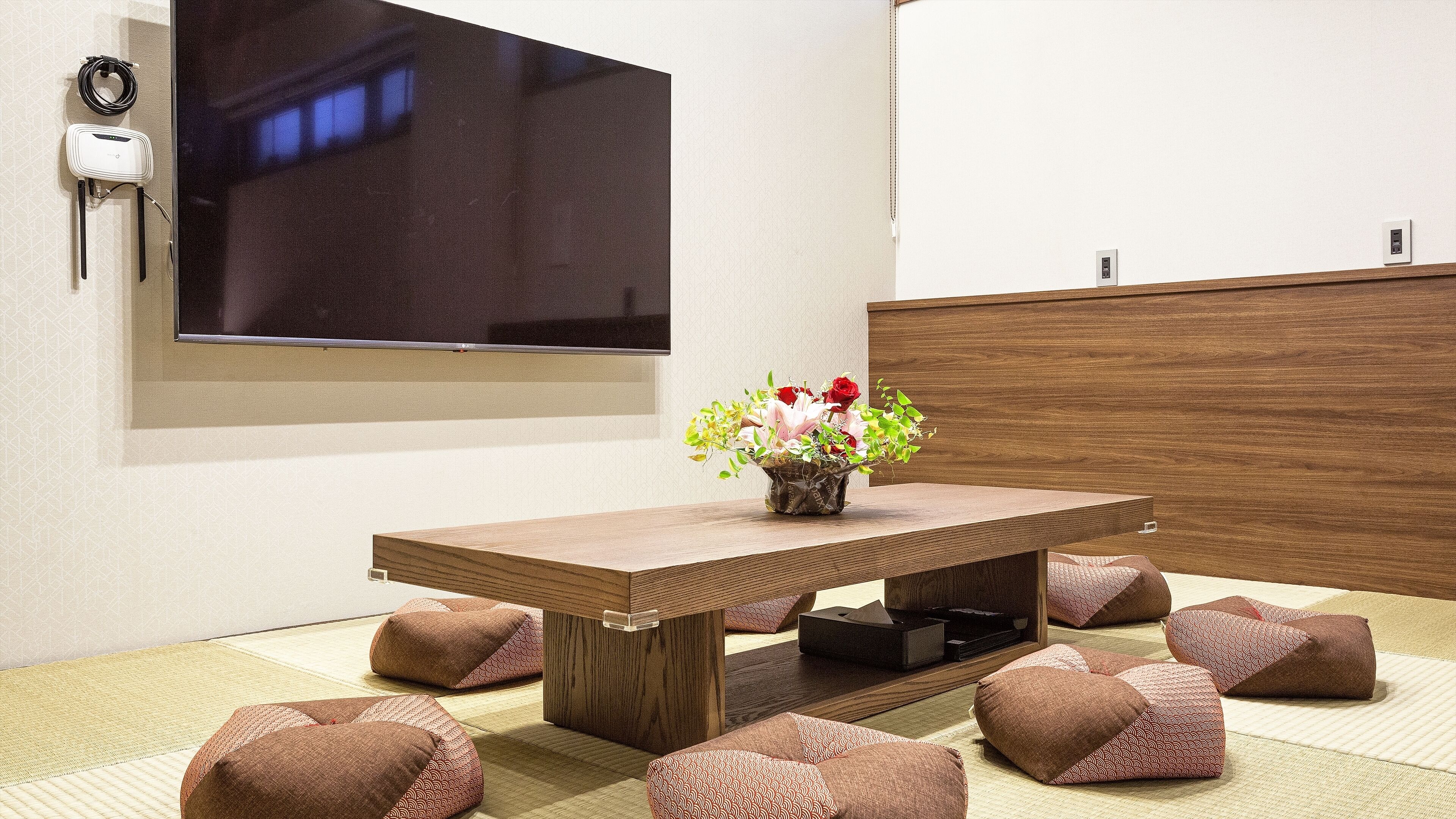 A modern dining area with a wooden table, a flat-screen TV mounted on the wall, and a vase with flowers.