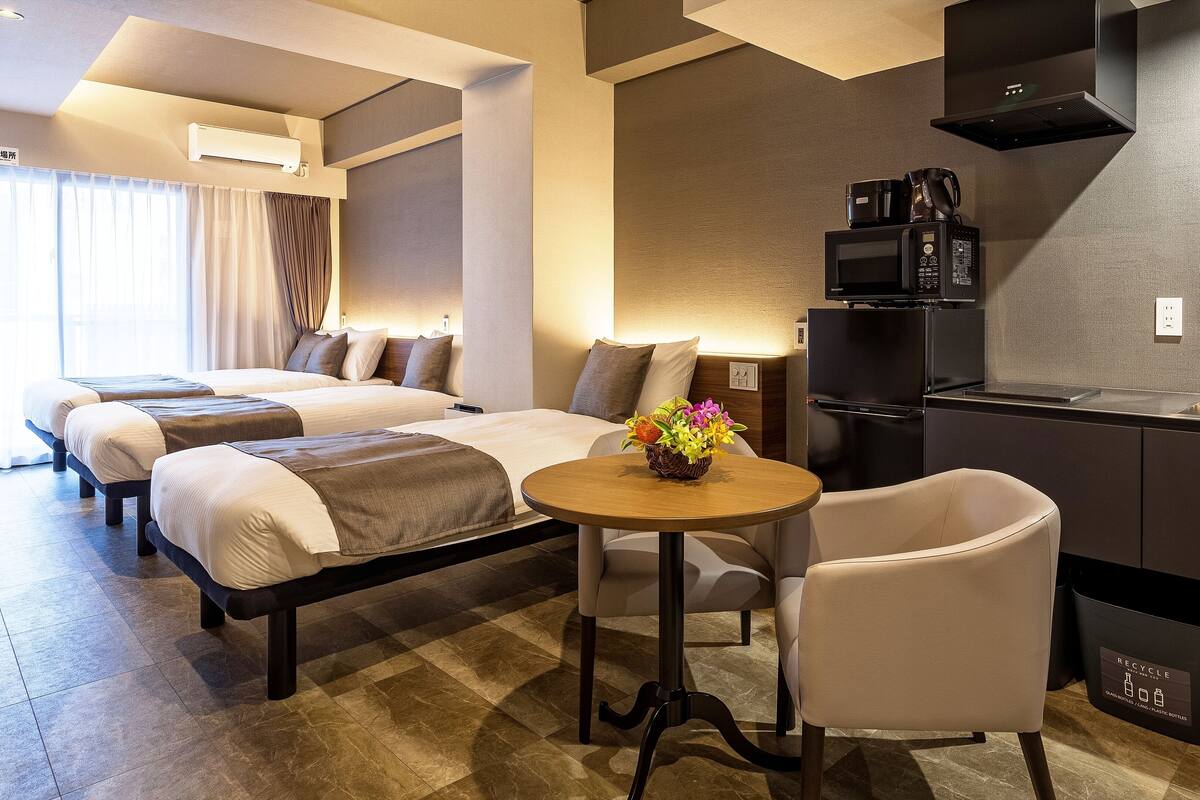 Une chambre d’hôtel avec deux lits, une petite table ronde ornée d’un vase de fleurs, un canapé, un coin cuisine avec un évier et une machine à café, et une poubelle de recyclage.