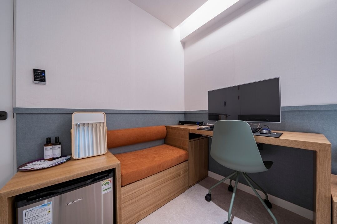 Habitación de hotel compacta con escritorio de madera, banca acolchada, refrigerador pequeño y televisor de pantalla plana.