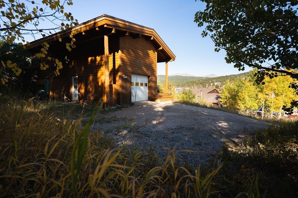 A wooden cabin with a gravel driveway, surrounded by trees and a distant view of a town.