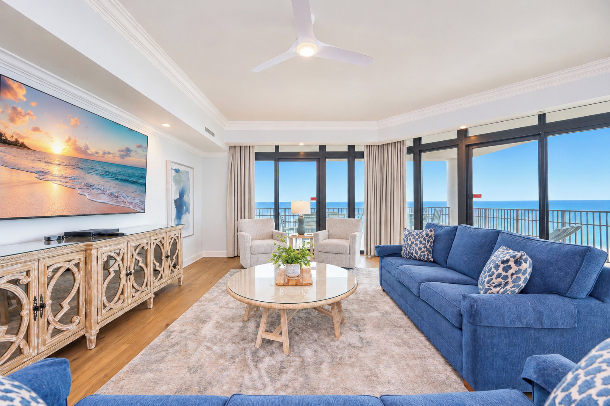 A spacious living room with a large TV, comfortable seating, a ceiling fan, and a view of the ocean.