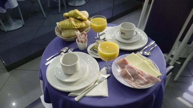 Une table de petit-déjeuner avec des assiettes, des couverts, un panier de pain et un verre de jus d’orange.