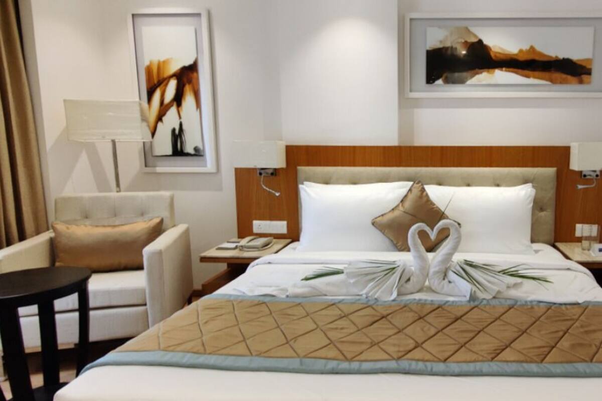 Une chambre d’hôtel avec un lit, une chaise, une table de chevet, une lampe et deux œuvres d’art encadrées accrochées au mur.