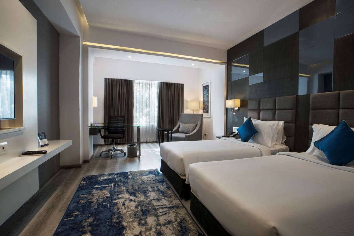 Une chambre d’hôtel avec deux lits, un bureau, une chaise, une télévision et une grande fenêtre avec des rideaux.