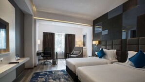 Habitación de hotel con dos camas, un escritorio, una silla, un televisor y un ventanal grande con cortinas.