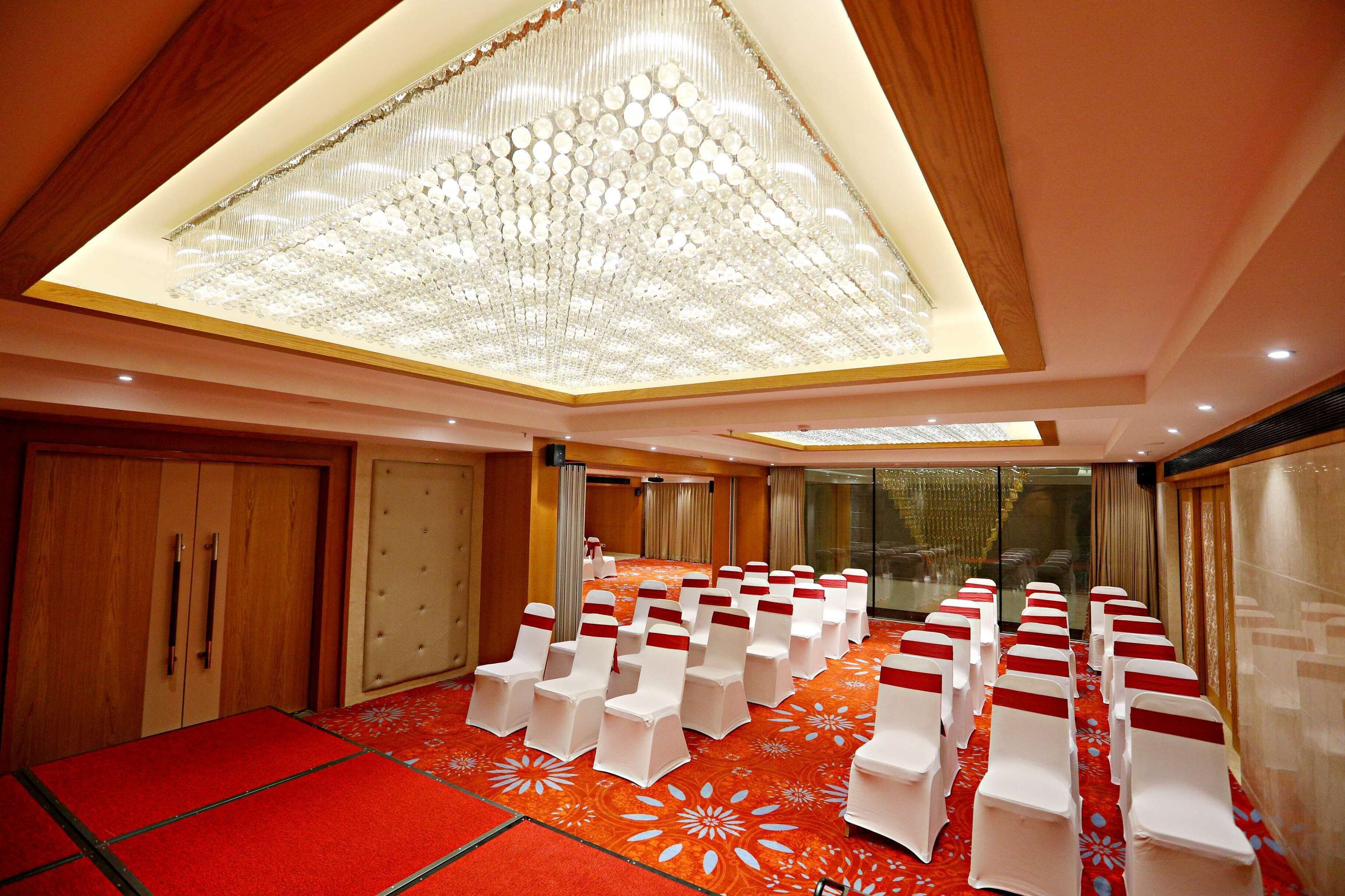 Un salón de conferencias con filas de sillas blancas adornadas con cintas rojas, una lámpara de araña grande y una alfombra roja con motivos florales blancos.