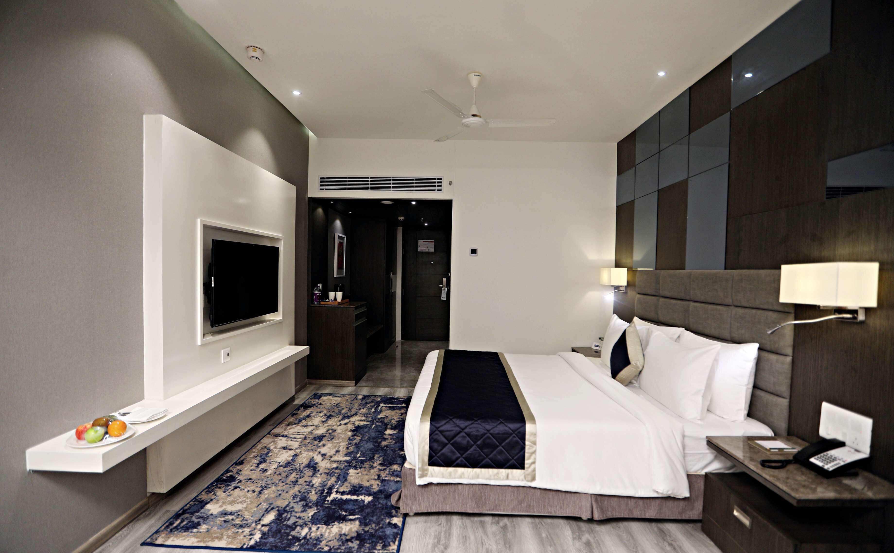 A modern hotel room with a flat-screen TV, a bed with white linens, a bedside table with a lamp, and a blue patterned rug.