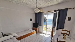 A room with two beds, a TV, a table, and a chair. There is a view of the sea through a sliding door.