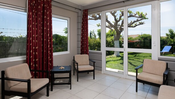 Ein sonnendurchflutetes Zimmer mit großen Fenstern, einem Sessel, einem kleinen Tisch und Blick auf einen Garten.