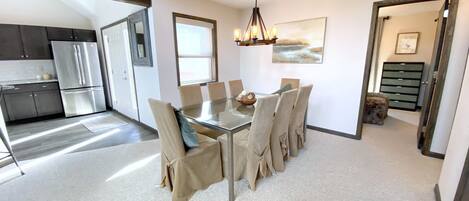 A modern kitchen with dark cabinets, a stainless steel refrigerator, and a dining area with a glass table and beige chairs.