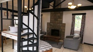 Una habitació amb una llar de pedra, taula de dinar amb caires, un sofà i una escala amb bigues al descobert.
