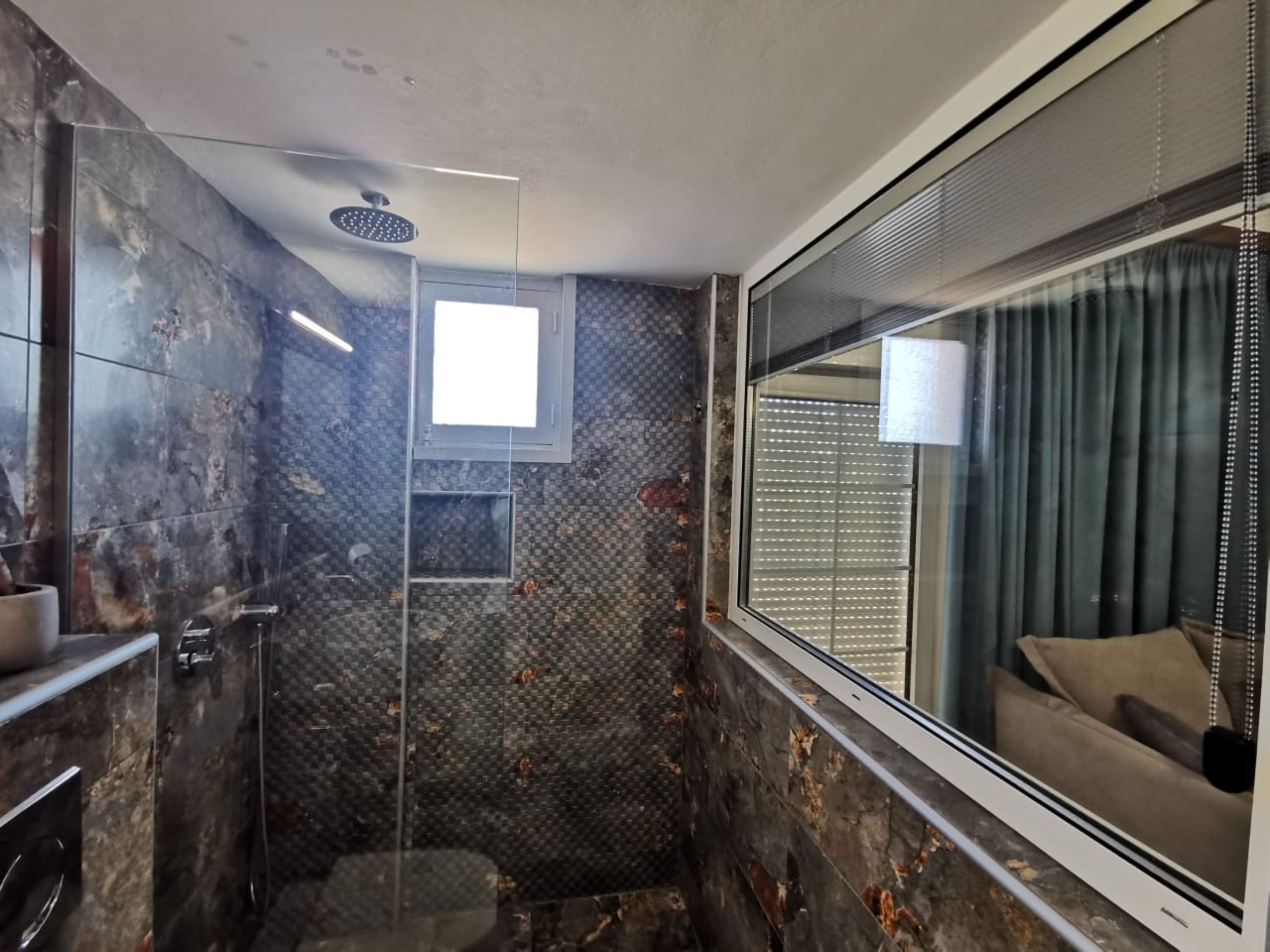 Een moderne badkamer met een douche, een raam met zonwering en uitzicht op een woonruimte.