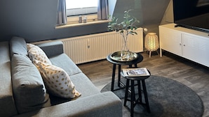 Una sala de estar moderna con un sofá gris, una mesita con una revista, una lámpara de pie y un televisor sobre un mueble blanco.