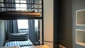 Una cama litera con estructura de madera y una cama individual debajo. Hay una mesita con un cojín frente a la cama. La habitación tiene una alfombra gris y una ventana con cortinas.