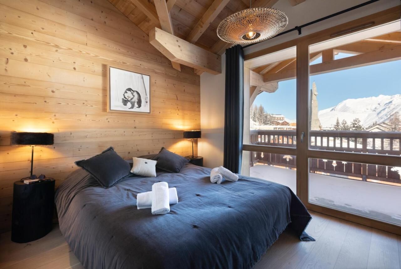 A wooden-paneled bedroom with a large bed, two bedside lamps, a framed picture, and a view of a snowy landscape through a balcony.