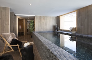 A modern indoor swimming pool with a tiled edge, a wooden chair with a fur throw, and a large window providing natural light. - TERRESENS - Neige et Soleil (Les Deux Alpes)
