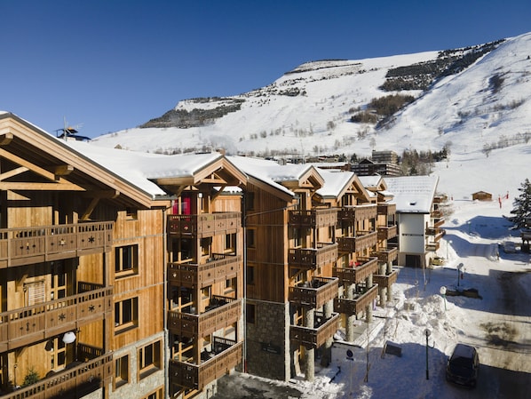 A row of wooden chalet-style buildings with balconies, set against a snowy mountain backdrop. - TERRESENS - Neige et Soleil (Les Deux Alpes)