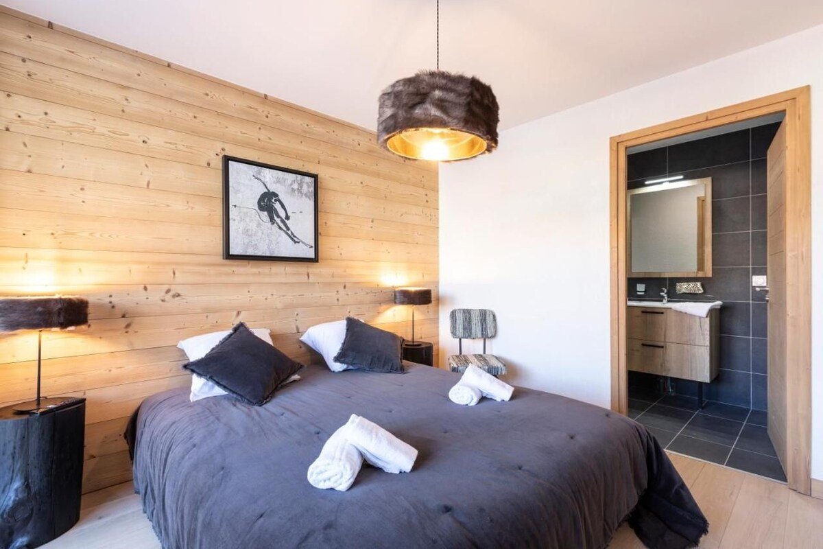 Une chambre avec un grand lit, des murs recouverts de panneaux en bois, un tableau encadré, un luminaire suspendu et une salle de bain visible à travers une porte ouverte.