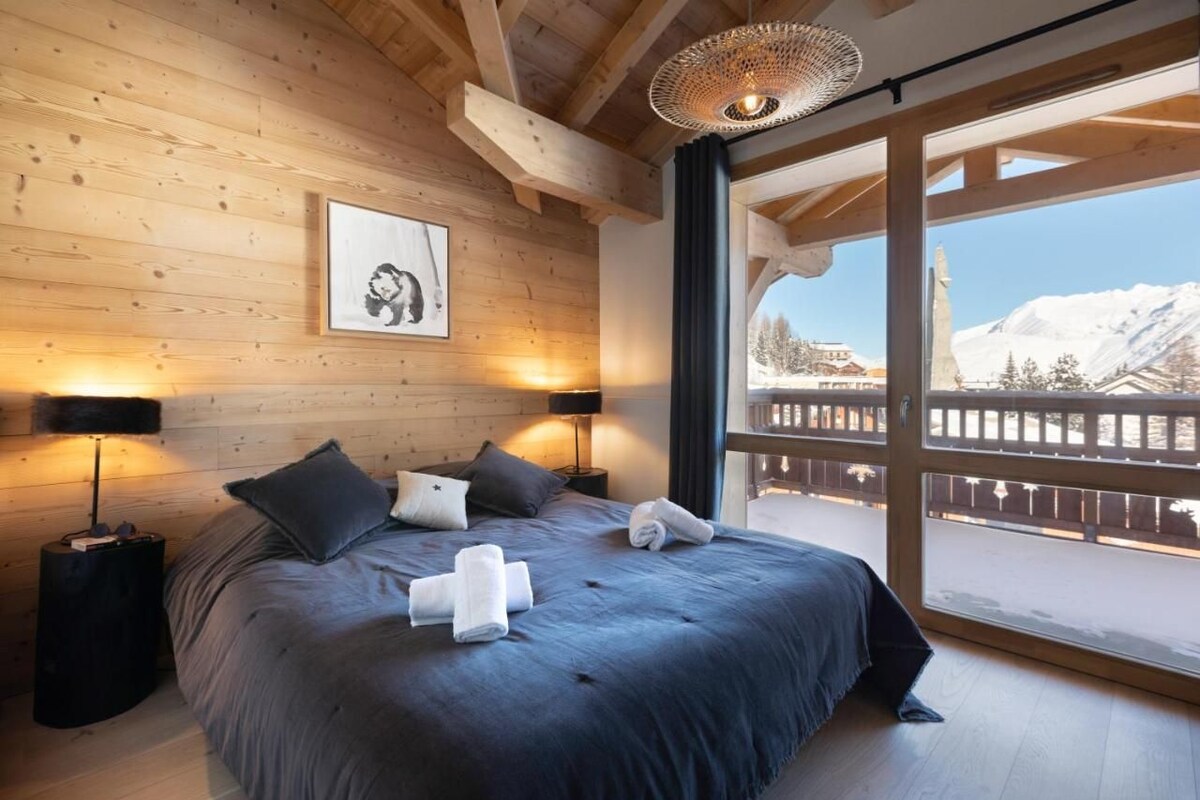 Quarto com painéis de madeira, uma cama grande, duas luminárias de cabeceira, um quadro e vista para uma paisagem nevada através de uma varanda.