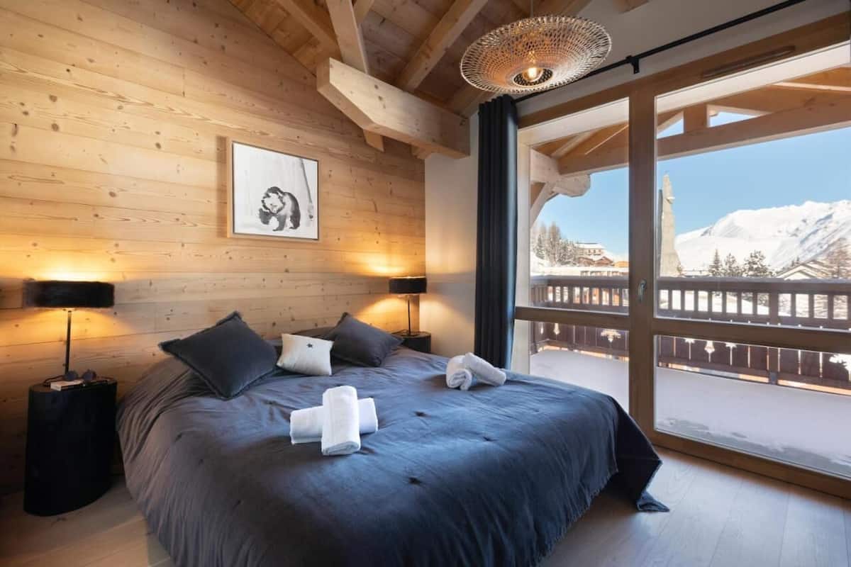Een slaapkamer met houten lambrisering, een groot bed, twee bedlampen, een ingelijst schilderij en uitzicht op een besneeerd landschap door een balkon.
