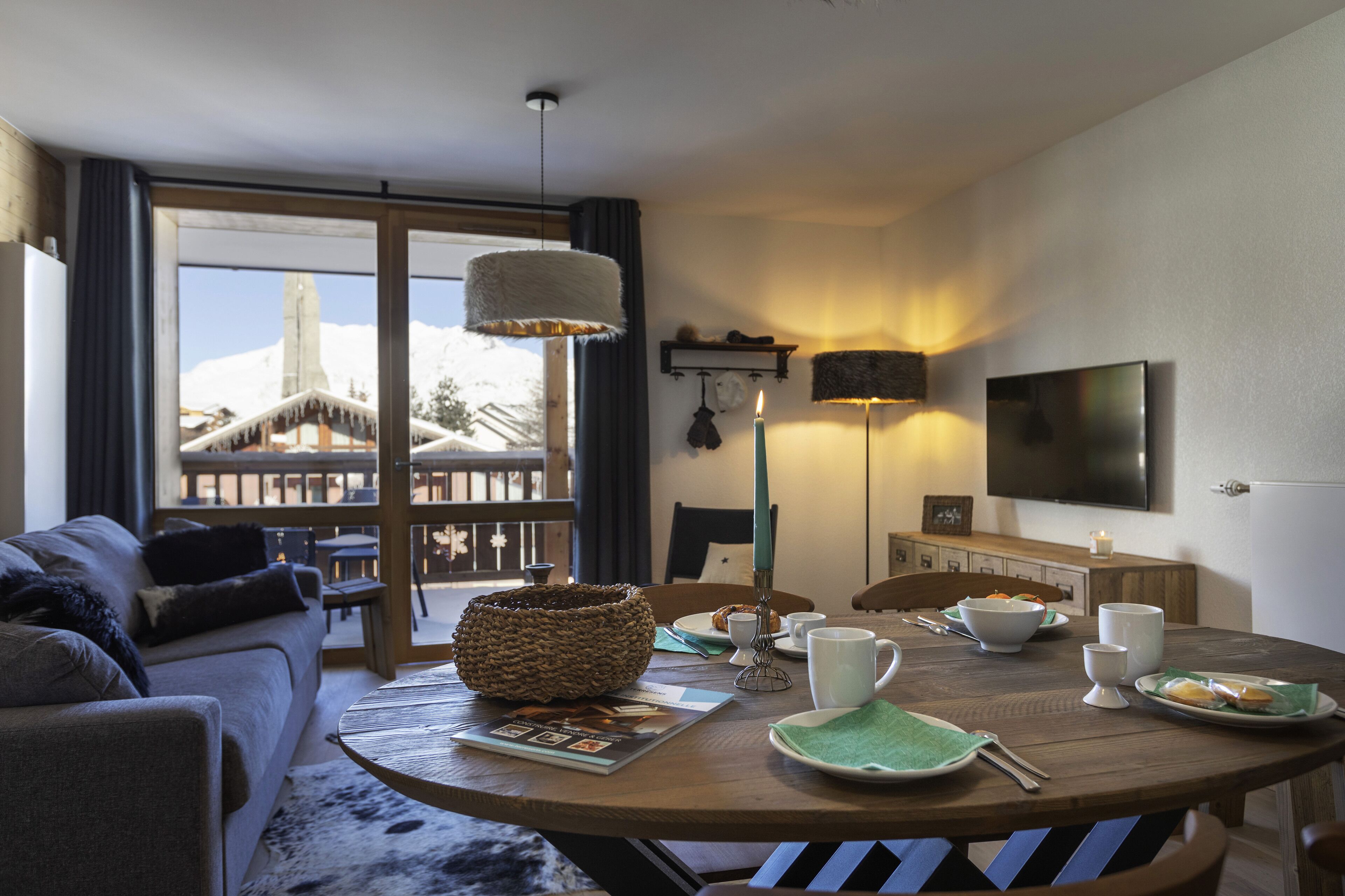 Uma sala de estar acolhedora com um sofá, uma mesa de jantar posta com pratos, xícaras e uma vela, e uma grande janela com vista para uma paisagem nevada.