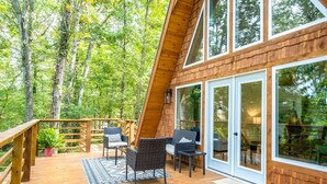 Une terrasse en bois avec deux fauteuils en osier et une table, entourée de portes et de fenêtres en verre, donnant sur une forêt.