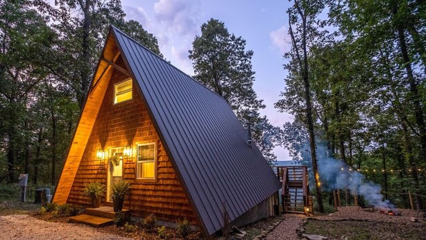 Une cabane en bois avec un toit en pente, un petit porche et une cheminée bien visible d’où s’échappe de la fumée.