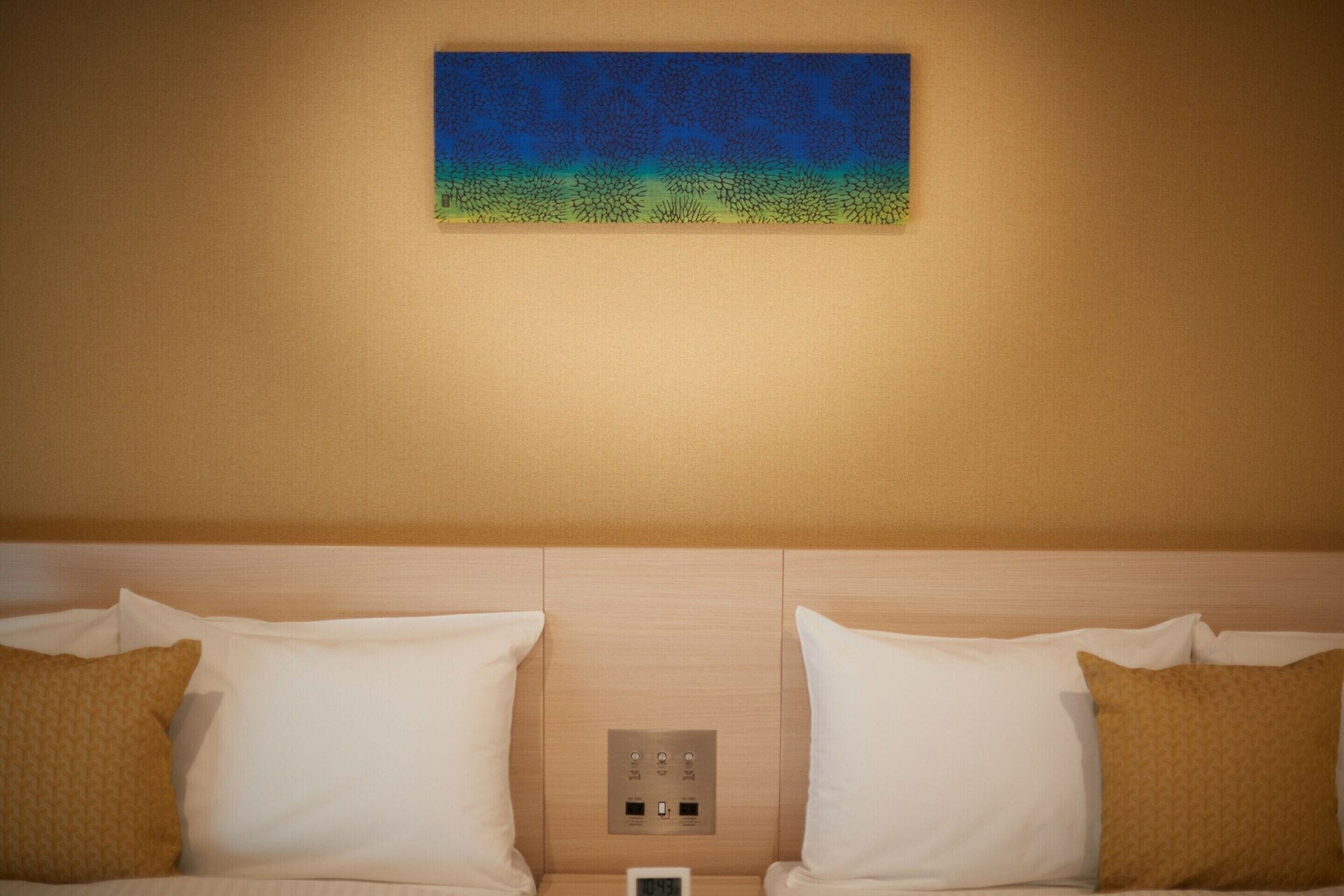 A hotel room with a bed, two pillows, a nightstand, a digital clock, and a wall-mounted artwork.