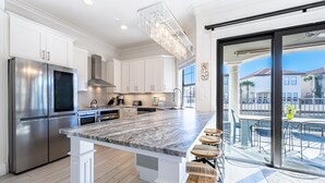 Une cuisine moderne avec des meubles blancs, des appareils en acier inoxydable, un îlot en marbre avec des tabourets de bar et une vue sur un patio aménagé avec du mobilier d’extérieur.