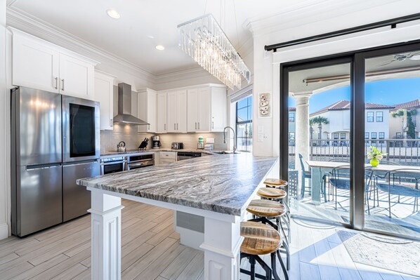 A modern kitchen with white cabinetry, stainless steel appliances, a marble island with bar stools, and a view of a patio with outdoor furniture.