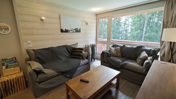 A cozy living room with two sofas, a wooden coffee table, and a large window with a view of trees. - Arcs 1800: Renovated 3BR 🏔️ 4 Crystals + Parking Incl. 🚗 - Transarc 100m (Bourg-Saint-Maurice)