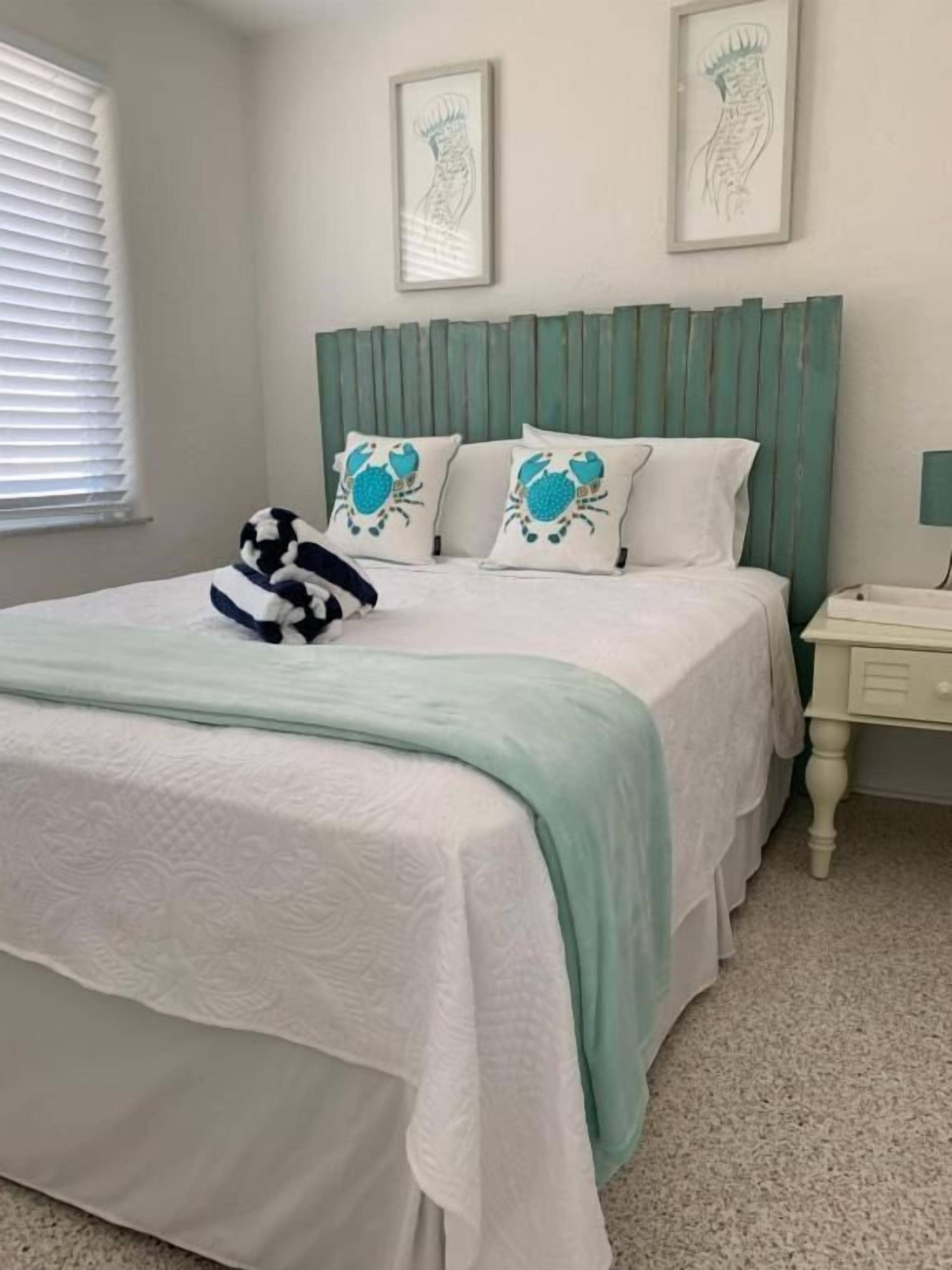 Un dormitorio ordenado con una cama, dos almohadas con motivos de cangrejo, un plaid a rayas y un cabecero.