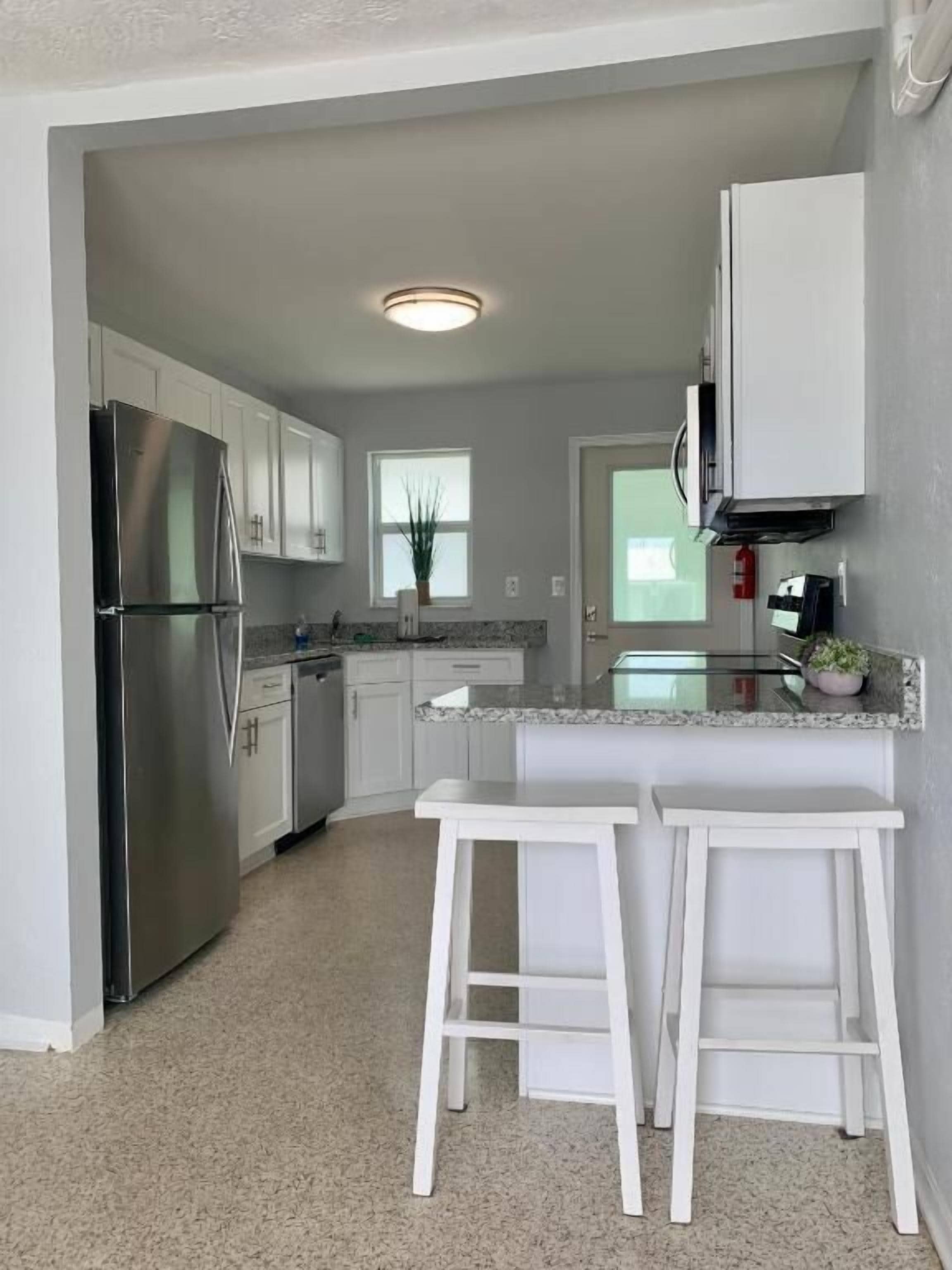 A modern kitchen with white cabinets, a stainless steel refrigerator, a granite countertop, and two white barstools.