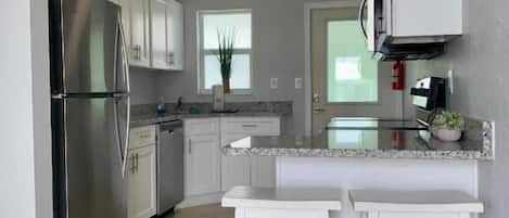 A modern kitchen with white cabinets, a stainless steel refrigerator, a granite countertop, and two white barstools.