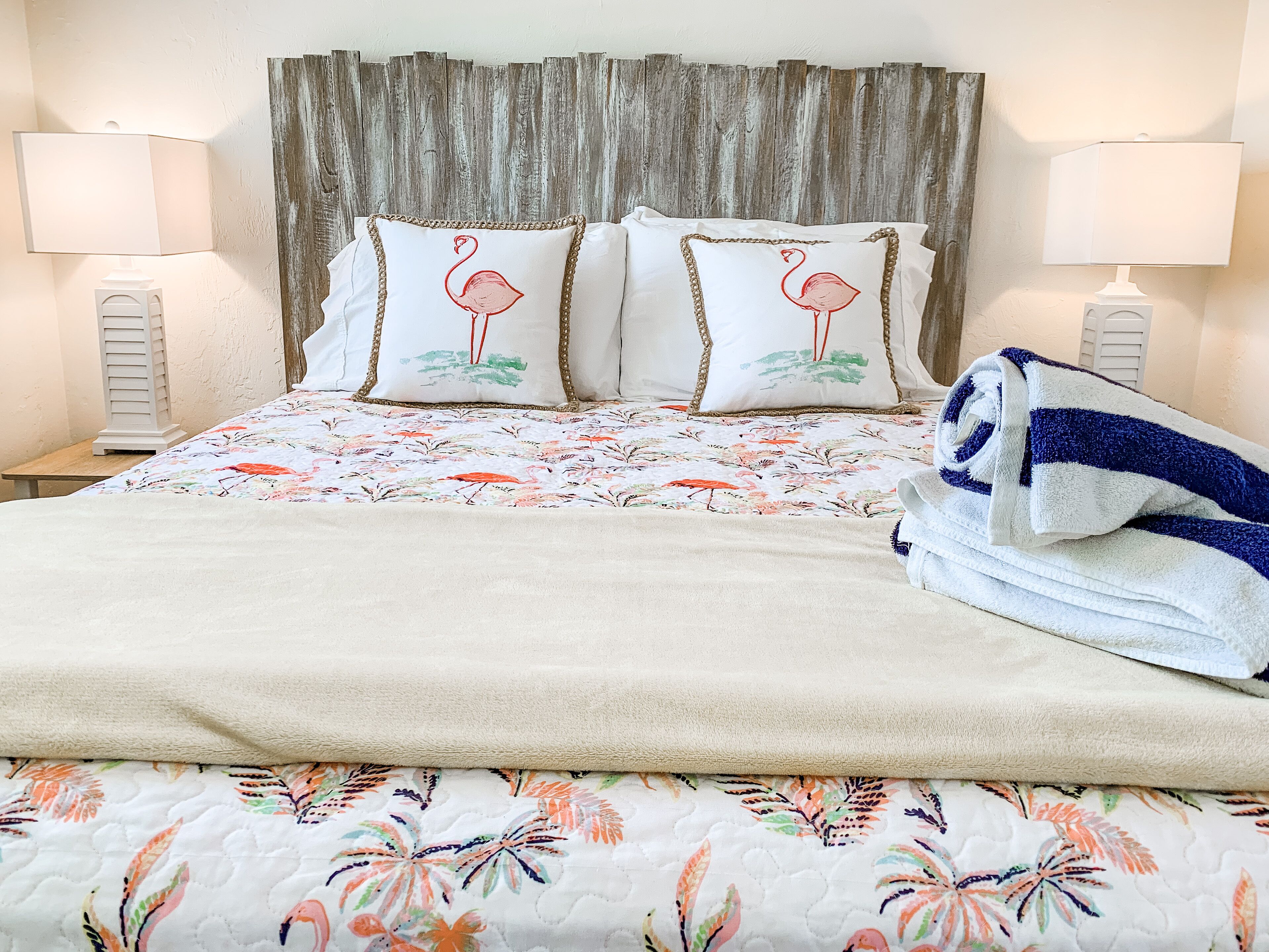 A bed with a floral bedspread, two pillows with flamingo designs, a folded towel, and a wooden headboard.