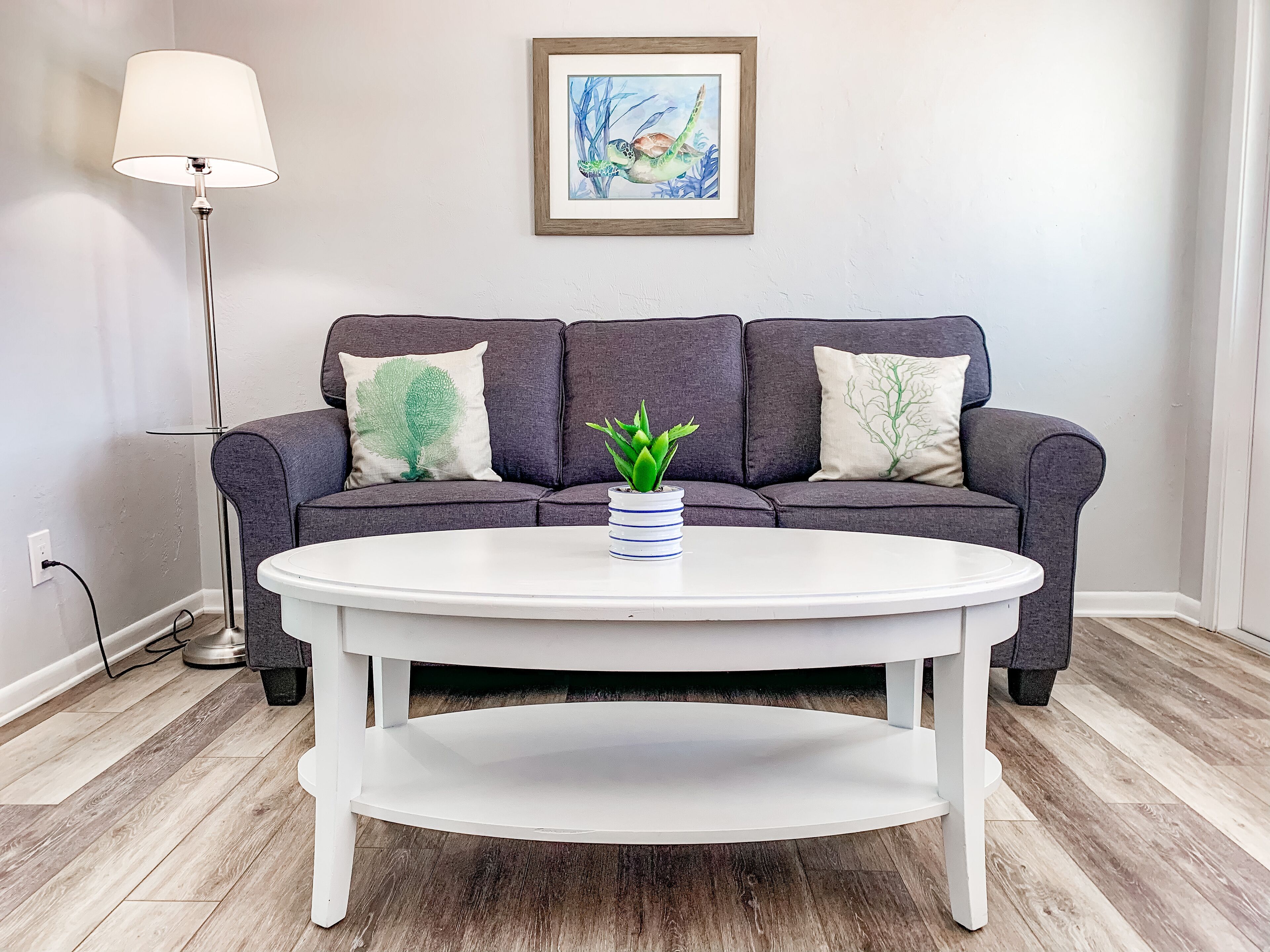 A modern living room with a grey sofa, two green decorative pillows, a white coffee table with a potted plant, and a framed artwork on the wall.