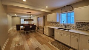 Une cuisine moderne avec des meubles blancs, un coin repas comprenant une table et des chaises, et une fenêtre équipée de stores.