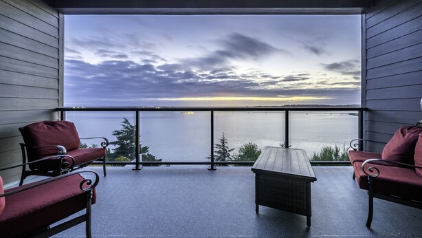 A balcony with two red chairs, a small table, and a view of a body of water and a sunset. - Luxurious Oregon Coast Townhouse with Mesmerizing Views! (Coos Bay)