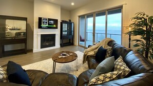 A living room with a fireplace, a television, a large window with a view, and a leather sofa. - Luxurious Oregon Coast Townhouse with Mesmerizing Views! (Coos Bay)