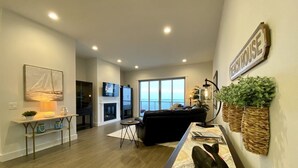 A modern living room with a sofa, a coffee table, and a TV. There is a fireplace, a window, and a sign that says "Beach House". - Luxurious Oregon Coast Townhouse with Mesmerizing Views! (Coos Bay)