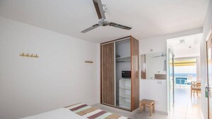 A bedroom with a bed, a ceiling fan, a wardrobe, a small table, and a view of the sea.