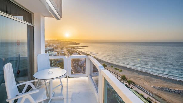 A balcony with a table and chairs, overlooking a beach and the ocean during sunset.