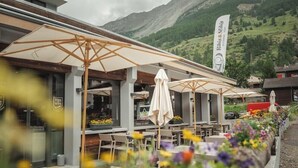 An outdoor seating area with umbrellas and tables, surrounded by flowers, with a mountain backdrop. - Matterhorn Inn (Taesch)