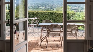 Een open deur die leidt naar een terras buiten, met een tafel en stoelen, een uitzicht op een bergachtig landschap en een haag op de achtergrond.