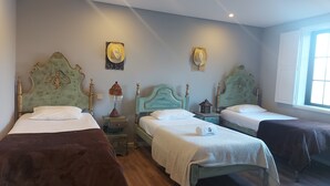 Habitación de hotel con tres camas individuales, cada una con un diseño de cabecera diferente. Mesitas de noche con lámparas y objetos decorativos. La habitación tiene una ventana con luz natural y vistas al exterior.